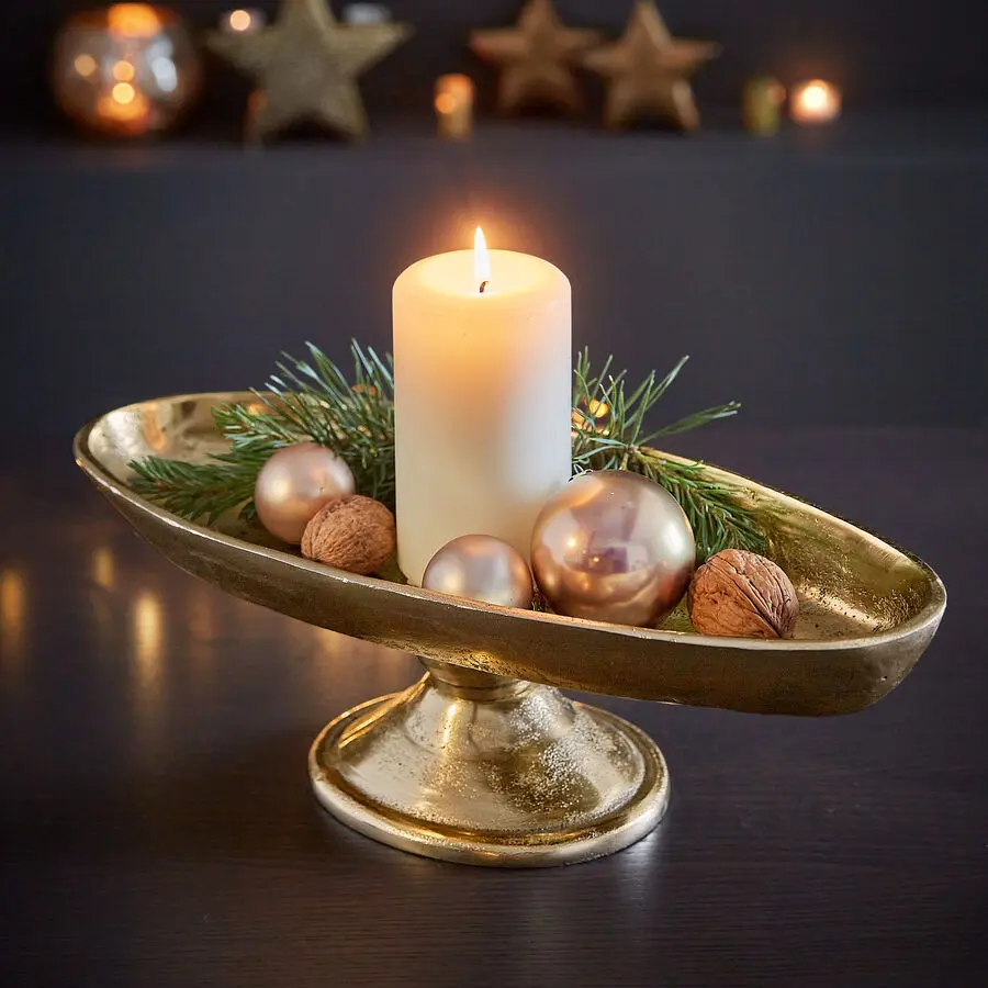Weiße Stumpenkerze in goldener Fußschale, dekoriert mit Tannengrün, glänzenden Kugeln und Walnüssen als Tischdeko