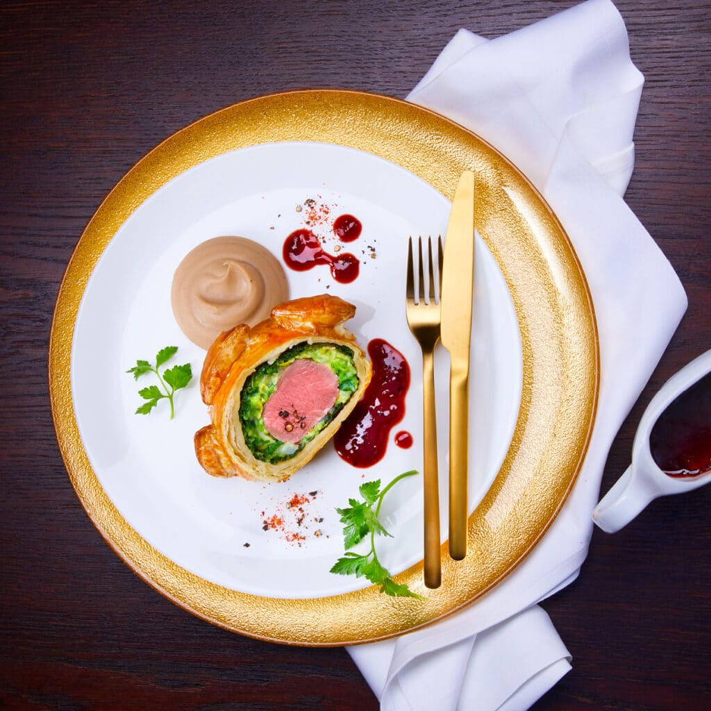 Festliches Rinderfilet Wellington im Blätterteig, serviert mit Gemüse, Sauce und cremigem Püree auf goldenem Teller