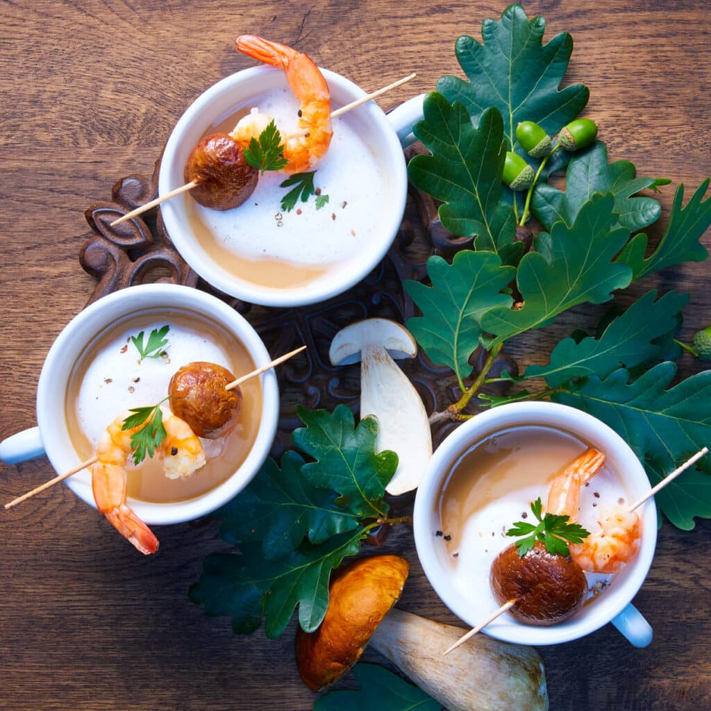 Cremige Suppe im Becher mit Garnelen und Maronen, dekoriert mit frischen Kräutern und Pilzen, festliche Vorspeise