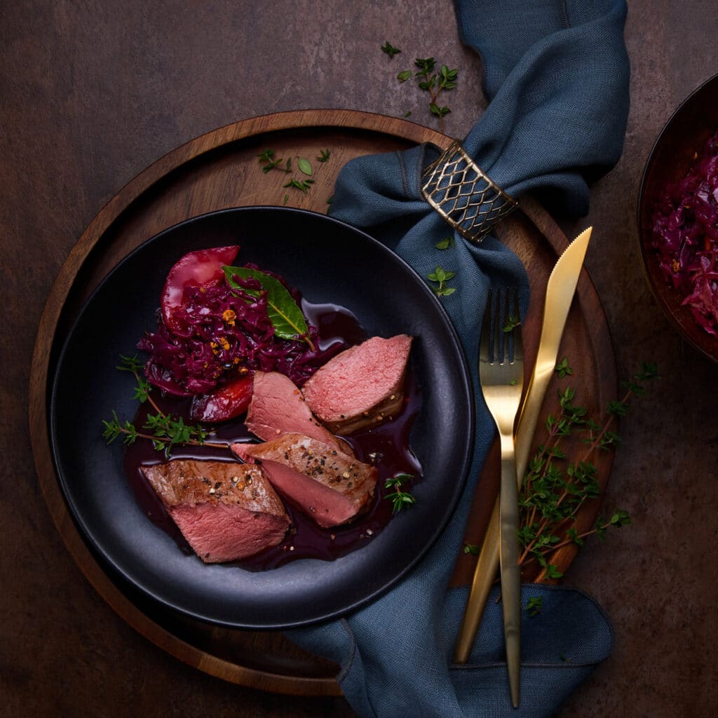 Rosa gebratener Rehrücken mit Rotkohl, Apfelscheiben und Jus auf schwarzem Teller