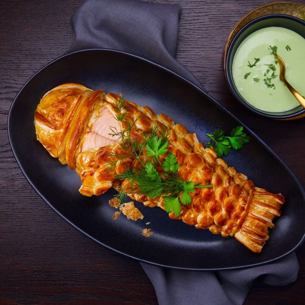 Ganzer Lachs im kunstvollen Blätterteigmantel mit grüner Sauce, perfekt für Weihnachten.