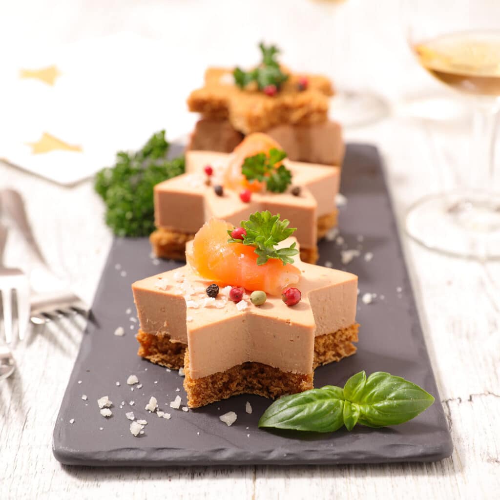 Weihnachtliche Foie gras Sterne auf knusprigem Brot, garniert mit Lachs, rosa Pfeffer, frischen Kräutern und Meersalz