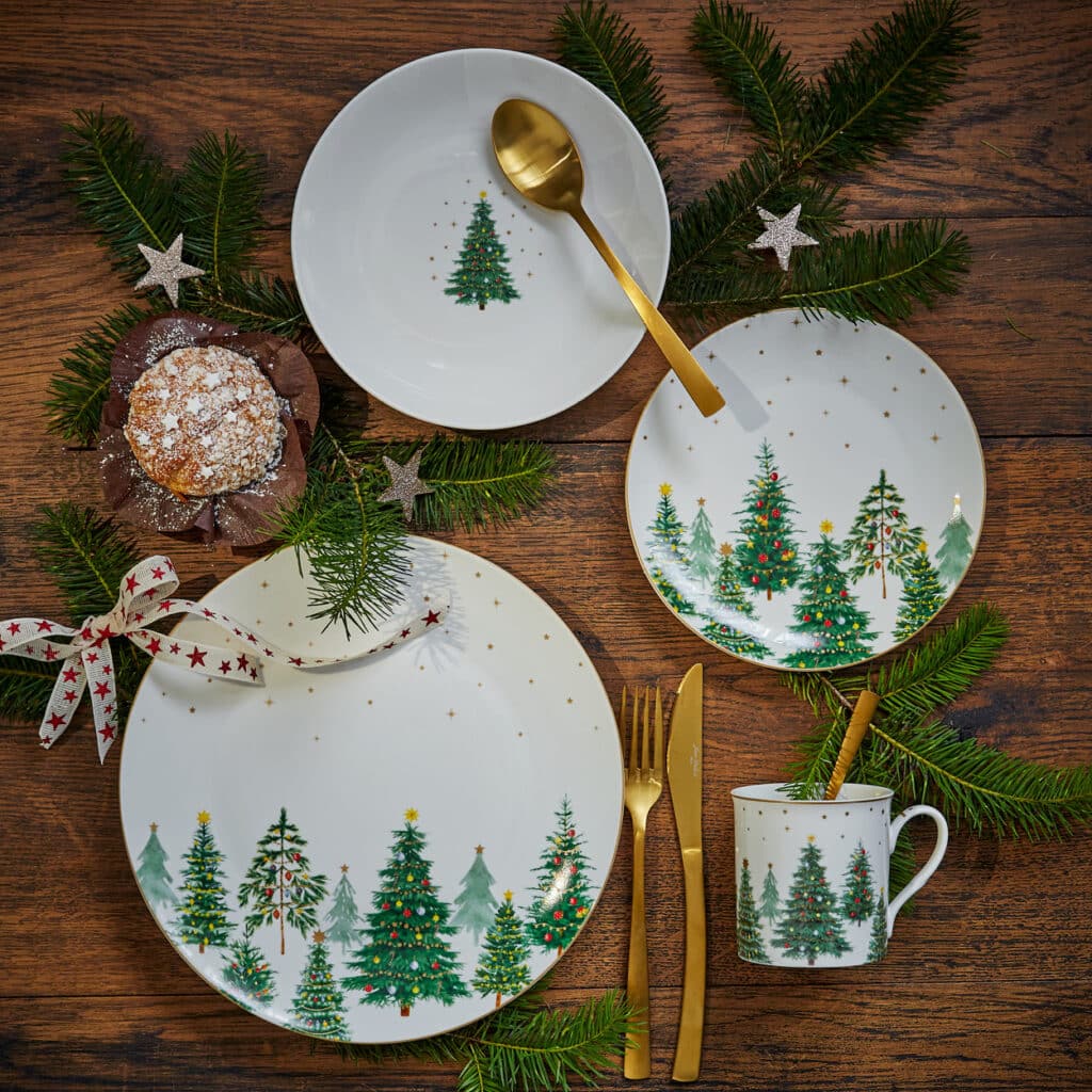 Weihnachtstafel mit Porzellan-Set im Tannenbaum-Dekor, goldfarbenes Besteck, festlich gedeckt mit Zweigen und Muffin