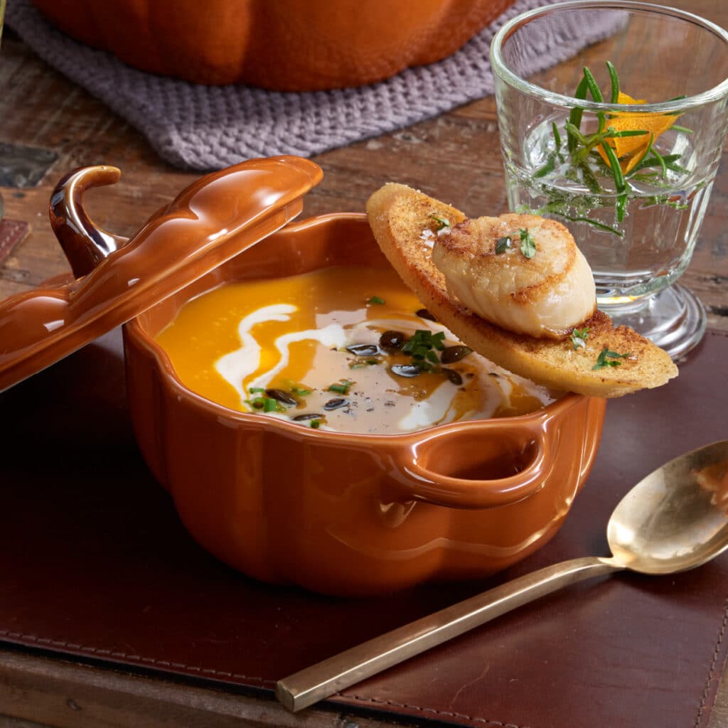 Kürbissuppe im festlichen Kürbisbecher, dekoriert mit Crostini und Jakobsmuschel, verfeinert mit Kräutern und Sahne