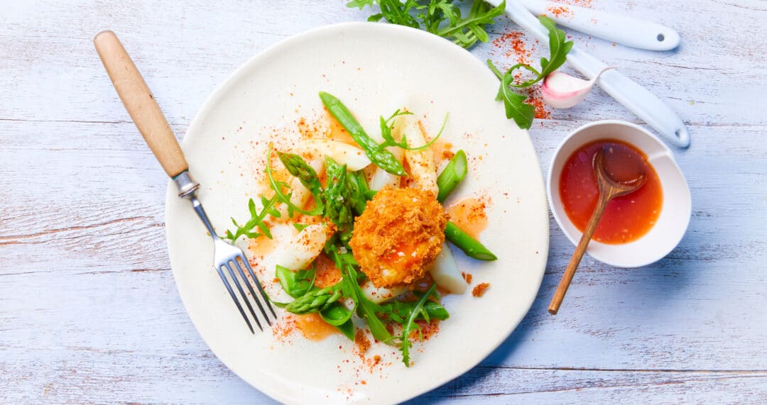 Teller mit Spargel, Salat und paniertem Ei, daneben Sch&auml;lchen mit Sauce und Besteck auf hellem Holztisch