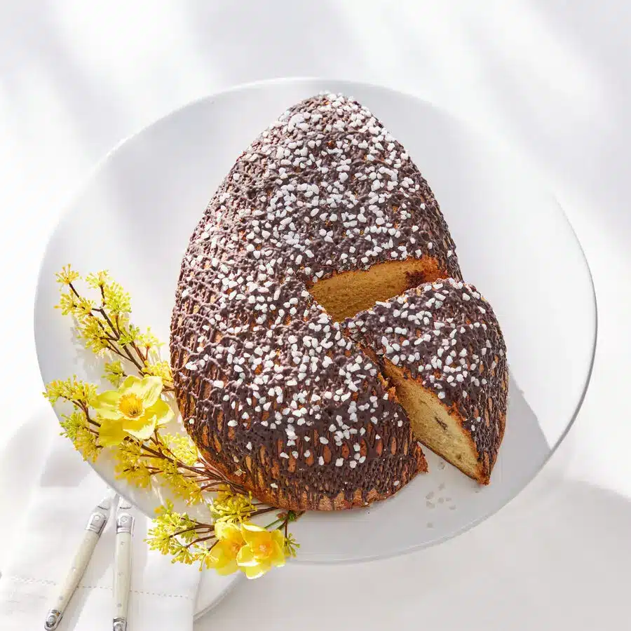 Oster-Panettone in Ei-Form mit Schokoladenglasur und Hagelzucker auf weißem Teller, angeschnittene Scheibe daneben
