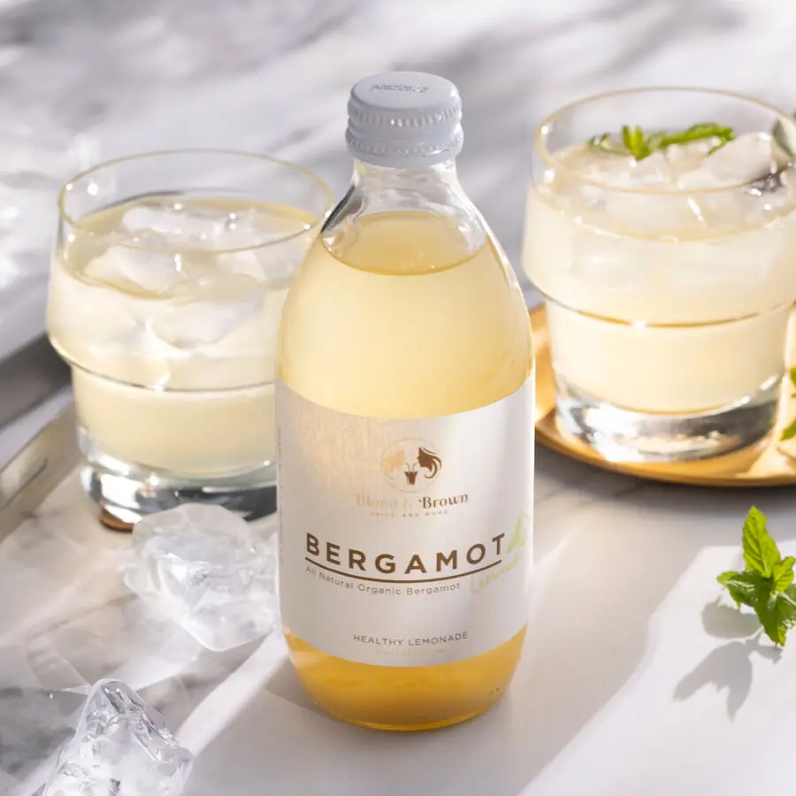 Flasche Bergamotte-Limonade mit Etikett, daneben zwei Gläser mit Eiswürfeln auf hellem Untergrund