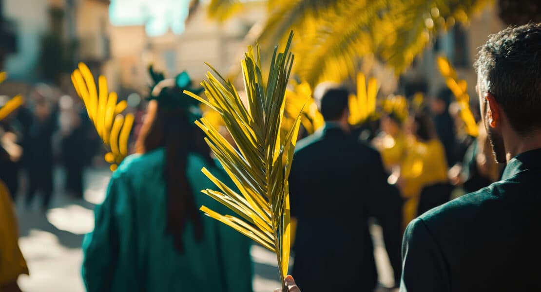Menschenmenge im Freien, eine Person hält einen grünen Palmzweig, weitere Palmblätter im Hintergrund