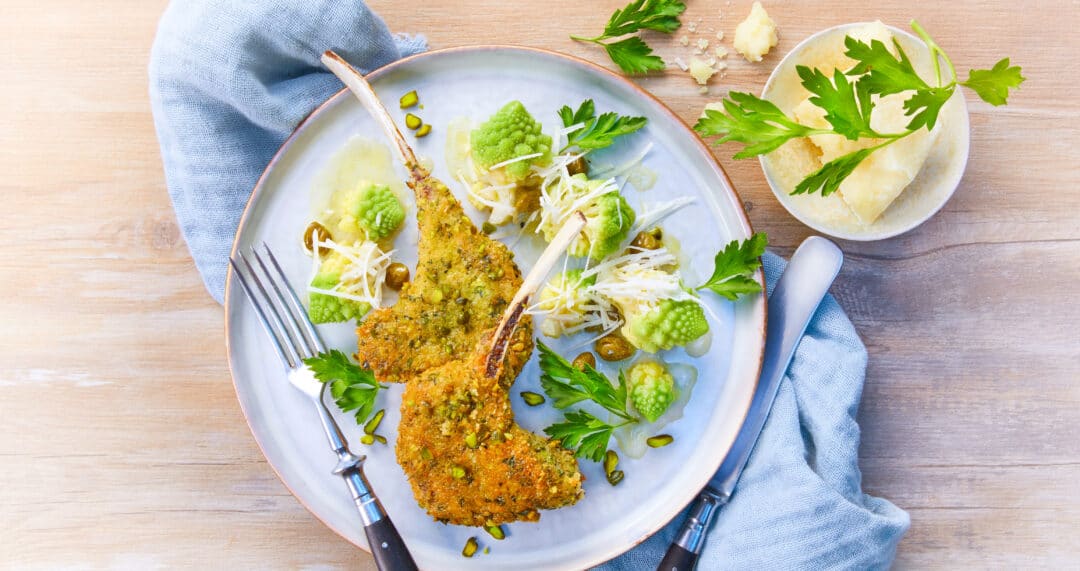 Panierte Lammkoteletts mit gr&uuml;nem Gem&uuml;se und gehobeltem K&auml;se auf hellem Teller, Besteck am Rand