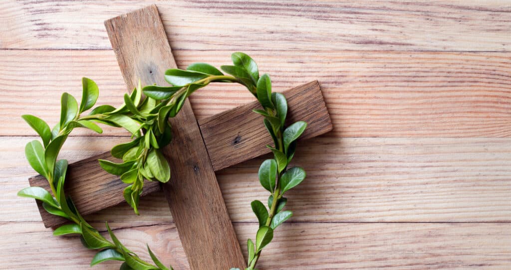 Holzkreuz auf Holzuntergrund, geschm&uuml;ckt mit gr&uuml;nen Zweigen in Kranzform