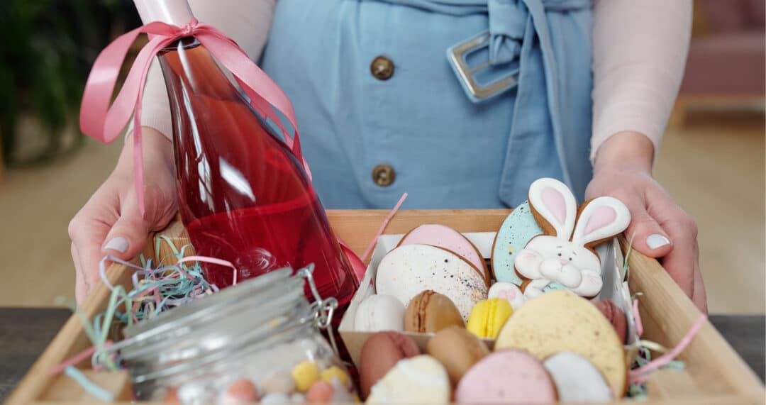 Person h&auml;lt eine Holzkiste mit S&uuml;&szlig;igkeiten, Macarons und einer Flasche mit rotem Getr&auml;nk