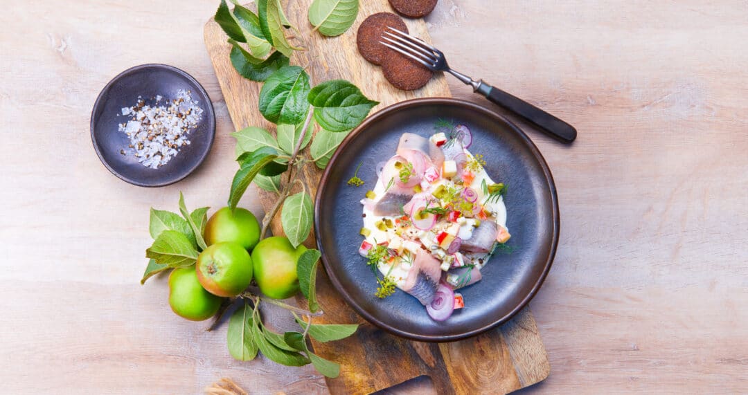 Fischst&uuml;cke mit Apfelw&uuml;rfeln, roten Zwiebeln und Kr&auml;utern in dunkler Schale, daneben gr&uuml;ne &Auml;pfel und Holzbrett