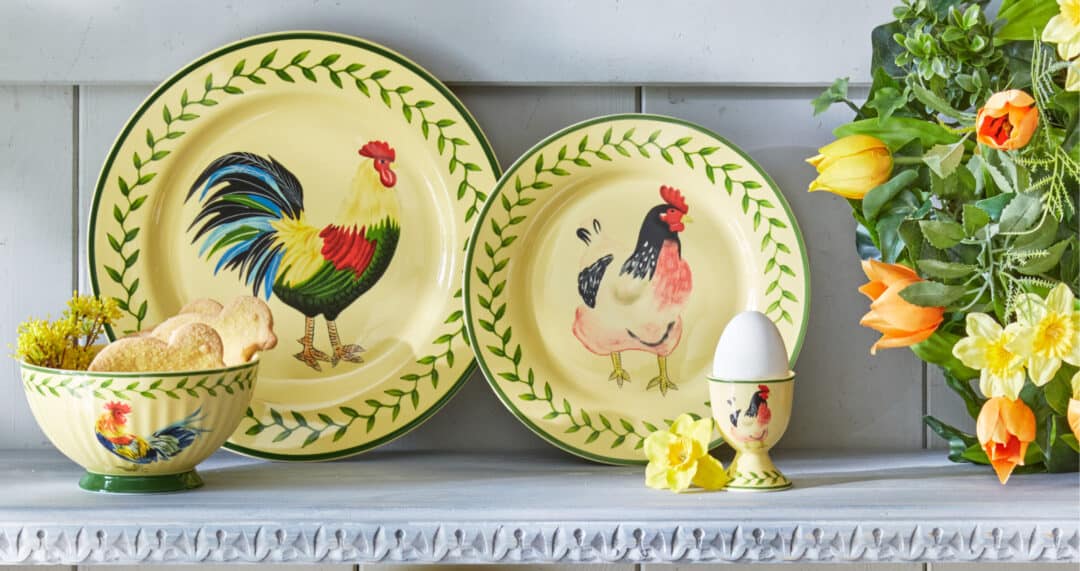 Fr&uuml;hlingshaftes Oster-Gedeck mit handbemalten Tellern im Hahn-und-Henne-Motiv, passender Schale und Eierbecher, dekoriert mit Fr&uuml;hlingsblumen auf hellem Regal.