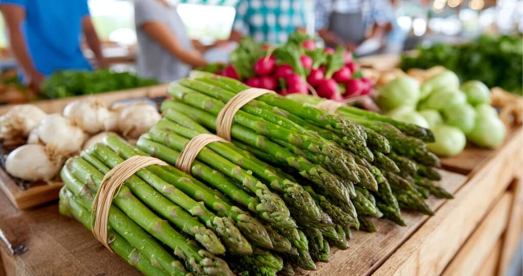 Frischer grüner Spargel gebündelt auf einem Marktstand, im Hintergrund Radieschen und Kohlrabi