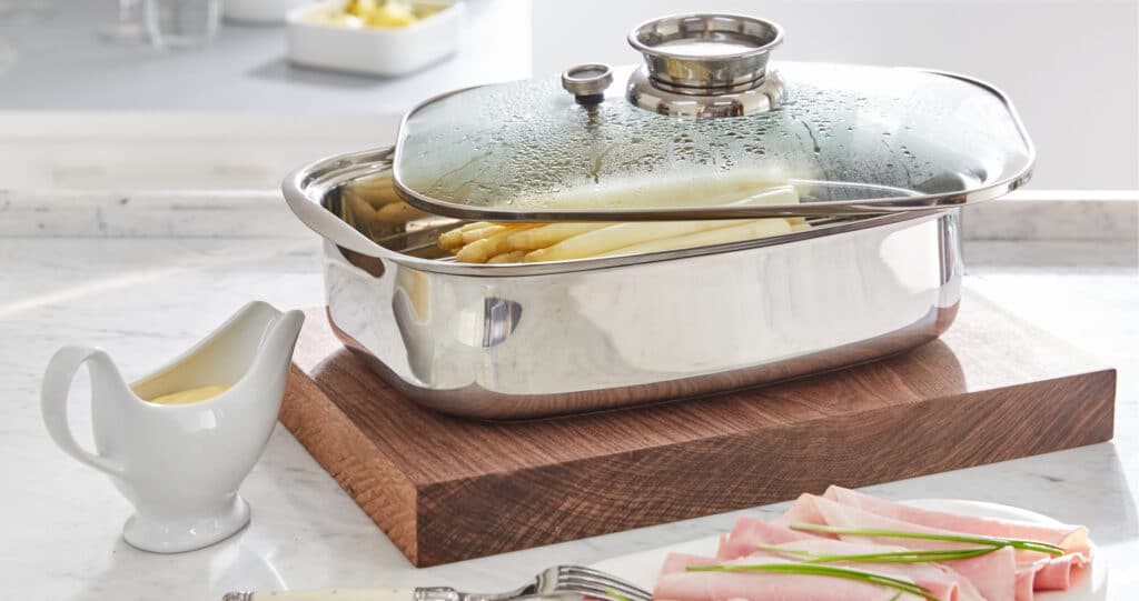 Weißer Spargel im Edelstahltopf mit Glasdeckel auf Holzbrett, dazu Sauce und gekochter Schinken